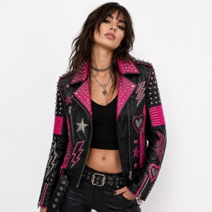 Puede incluir: Una chaqueta de moto de cuero negro con paneles de cuero rosa vibrante, tachuelas de metal plateado y parches bordados en forma de estrellas, corazones y rayos. La chaqueta incluye un cuello de solapa, bolsillos con cremallera y un cinturón.