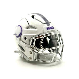 May include: A white miniature football helmet with a chrome face mask. The helmet has purple accents and the word "VIKINGS" printed on the front. The helmet is a collectible item.