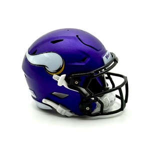 May include: A purple football helmet with a white and gold logo on the side. The helmet has a black face mask and is set against a white background. The word "Vikings" is visible on the helmet.