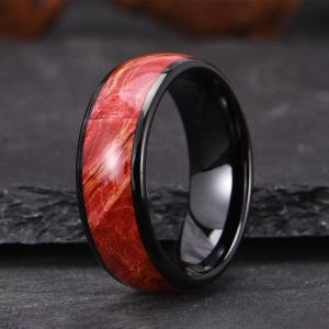 Può includere: Un anello caratterizzato da una fascia nera lucida con un ampio inserto in legno rosso venato. L'inserto in legno presenta motivi naturali a vortice in tonalità di rosso cremisi intenso e arancione brillante, in contrasto con i bordi in metallo nero.