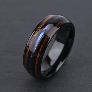 May include: A polished black ring featuring a central inlay of brown wood grain and a thin, iridescent blue abalone shell strip running through the middle. The band has a smooth, reflective finish and a rounded profile with a dark, sleek appearance.