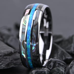May include: A polished dark metal ring featuring a central inlay of bright blue crushed opal flanked by two parallel strips of iridescent abalone shell. The band has a smooth, reflective finish with distinct metallic edges framing the inlays.