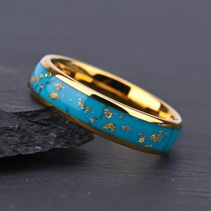 May include: A gold-toned ring featuring a bright turquoise inlay that is speckled with small, irregular gold-colored flakes throughout the band. The ring has a polished, reflective gold interior and a smooth, rounded outer surface.
