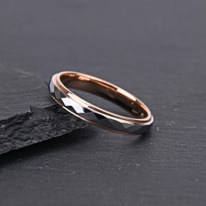 May include: A ring featuring a faceted dark gray outer band with a polished rose gold interior. The exterior surface has a geometric, multi-faceted design that reflects light, while the inner band is smooth and metallic in a warm rose gold tone.