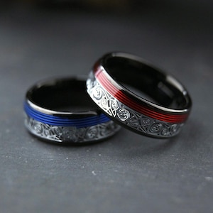 Op de afbeelding: Twee ringen van zwart metaal met ingewikkelde zilverkleurige krulpatronen. De ene ring heeft een felblauwe gegroefde middenstreep, terwijl de andere ring een opvallende rode gegroefde middenstreep heeft, beide met een gepolijste afwerking.