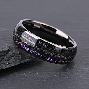 Puede incluir: Un anillo de banda negra con acabado pulido y una incrustación central de escamas trituradas iridiscentes en tonos morados, azules y plateados que crean un efecto brillante y multicolor sobre la superficie metálica oscura y lisa.