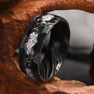 May include: A black ring featuring a textured, dark metallic band with a central inlay of shimmering silver-colored flakes. The surface of the ring displays a distinct, fine-lined pattern across the dark metal, contrasting with the bright, irregular silver accents embedded within the center of the band.