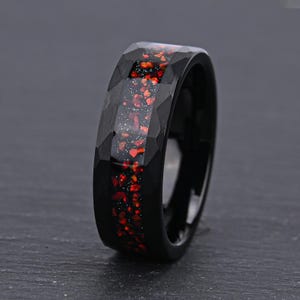Può includere: Un anello nero con una superficie esterna sfaccettata e un intarsio centrale caratterizzato da uno sfondo scuro con piccole macchie irregolari rosse e bianche, che creano un aspetto maculato e multicolore al centro della fascia.