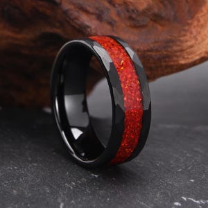 May include: A black ring with a faceted outer edge and a wide, central inlay of crushed red material. The band features a smooth, polished interior surface and a dark, reflective finish that contrasts with the vibrant, textured red inlay.