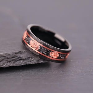 May include: A black ring featuring a faceted outer band with a central inlay of dark, textured material interspersed with irregular, metallic copper-coloured flakes. The ring has a smooth, polished interior and a dark, matte-finished exterior surface.