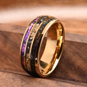 May include: A gold-toned ring featuring a wide, polished band with a central inlay. The inlay consists of three distinct, textured channels filled with iridescent purple, shimmering gold, and dark green crushed mineral fragments.