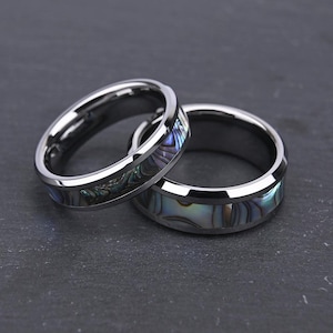 May include: Two polished silver-toned metal rings featuring a central inlay of iridescent abalone shell. The rings have a smooth, beveled edge design, showcasing the natural swirling patterns of blue, green, and purple colors within the shell material.
