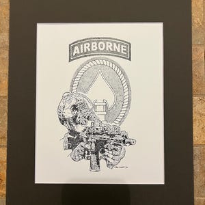 May include: Black and white illustration of a soldier holding a rifle, with the word "AIRBORNE" above. The artwork is on white paper, matted in black, and framed.