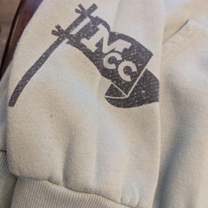 Puede incluir: Primer plano de una manga de sudadera verde claro. Un gr&aacute;fico de bandera gris oscuro con las letras "NCC" est&aacute; impreso en la manga. El pu&ntilde;o es acanalado y la tela parece suave y ligeramente desgastada.