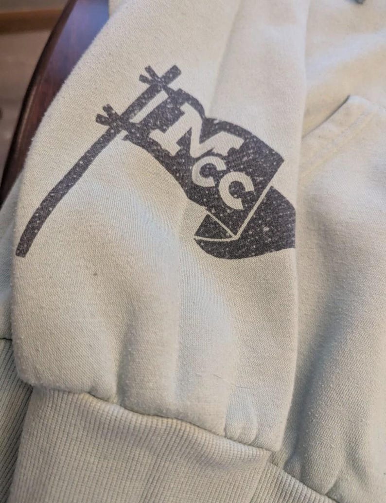 Puede incluir: Primer plano de una manga de sudadera verde claro. Un gr&aacute;fico de bandera gris oscuro con las letras "NCC" est&aacute; impreso en la manga. El pu&ntilde;o es acanalado y la tela parece suave y ligeramente desgastada.