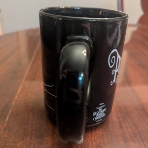 May include: A black ceramic mug featuring a white graphic design and the text Disney Tim Burton's The Nightmare Before Christmas. The mug has a curved handle on the side and a smooth, glossy finish. The text is printed in a stylised white font.