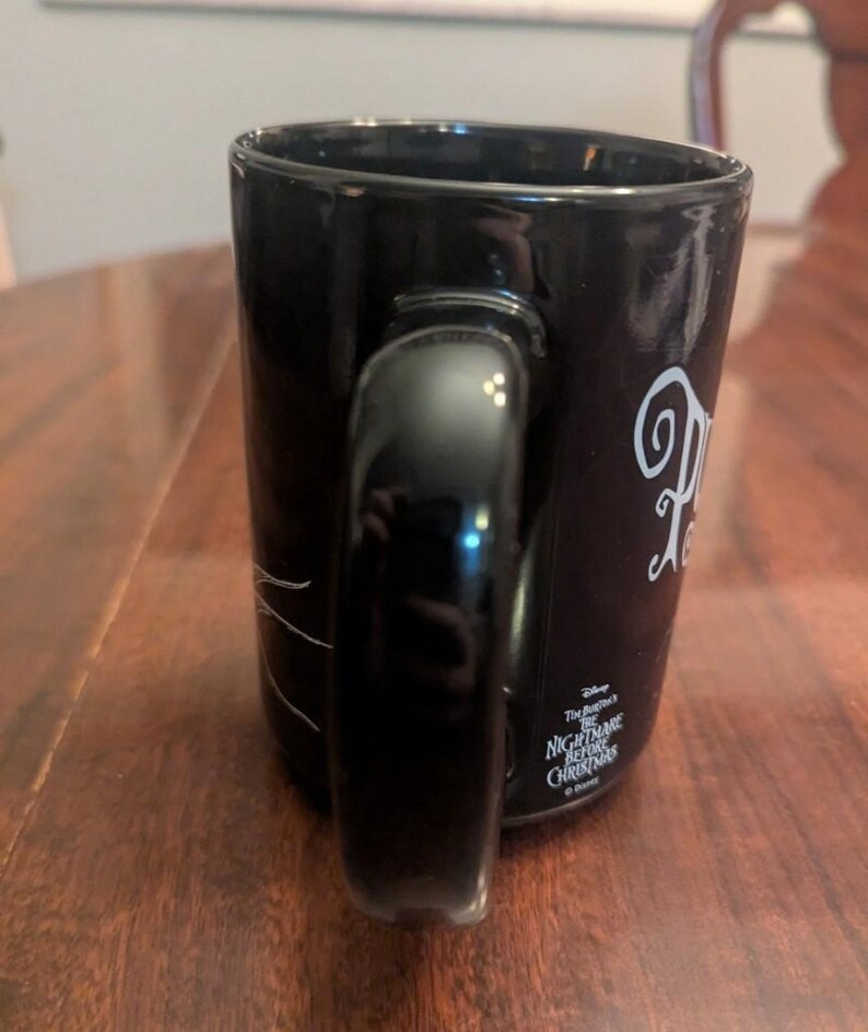 May include: A black ceramic mug featuring a white graphic design and the text Disney Tim Burton's The Nightmare Before Christmas. The mug has a curved handle on the side and a smooth, glossy finish. The text is printed in a stylised white font.