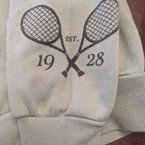 Puede incluir: Manga de sudadera verde claro con pu&ntilde;o acanalado. Dos raquetas de tenis cruzadas est&aacute;n impresas en gris oscuro, con el texto "EST. 1928" arriba y abajo. La sudadera est&aacute; hecha de un material suave.