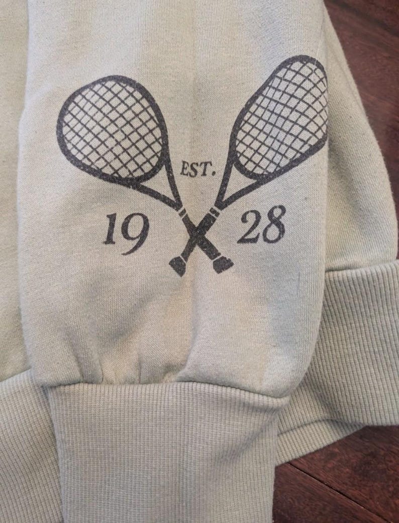 Puede incluir: Manga de sudadera verde claro con pu&ntilde;o acanalado. Dos raquetas de tenis cruzadas est&aacute;n impresas en gris oscuro, con el texto "EST. 1928" arriba y abajo. La sudadera est&aacute; hecha de un material suave.