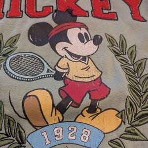 Puede incluir: Sudadera verde claro con un dise&ntilde;o vintage de Mickey Mouse jugando al tenis. El dise&ntilde;o incluye la palabra "MICKEY" en rojo, el a&ntilde;o "1928" y hojas de laurel. Mickey lleva una camisa amarilla, pantalones cortos rojos y una diadema roja.