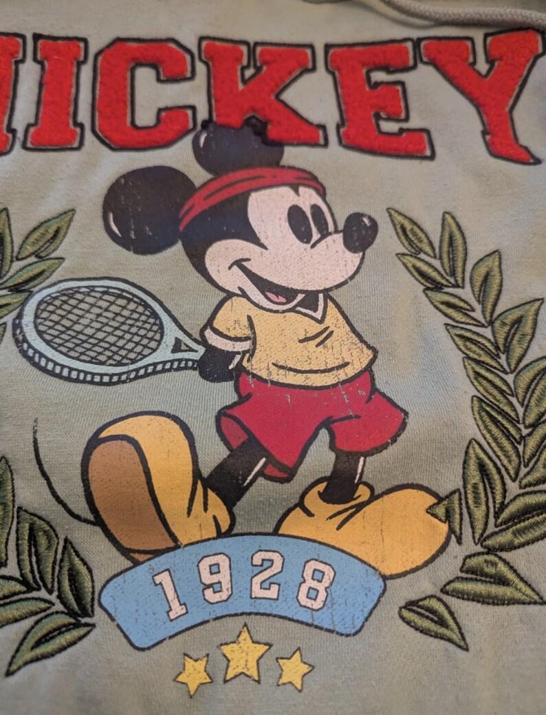 Puede incluir: Sudadera verde claro con un dise&ntilde;o vintage de Mickey Mouse jugando al tenis. El dise&ntilde;o incluye la palabra "MICKEY" en rojo, el a&ntilde;o "1928" y hojas de laurel. Mickey lleva una camisa amarilla, pantalones cortos rojos y una diadema roja.