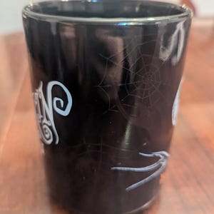 May include: A black cylindrical mug featuring a glossy finish with a white spiderweb pattern printed on the exterior. The design includes stylised white lettering and a grey spider graphic, creating a dark aesthetic on the smooth ceramic surface.