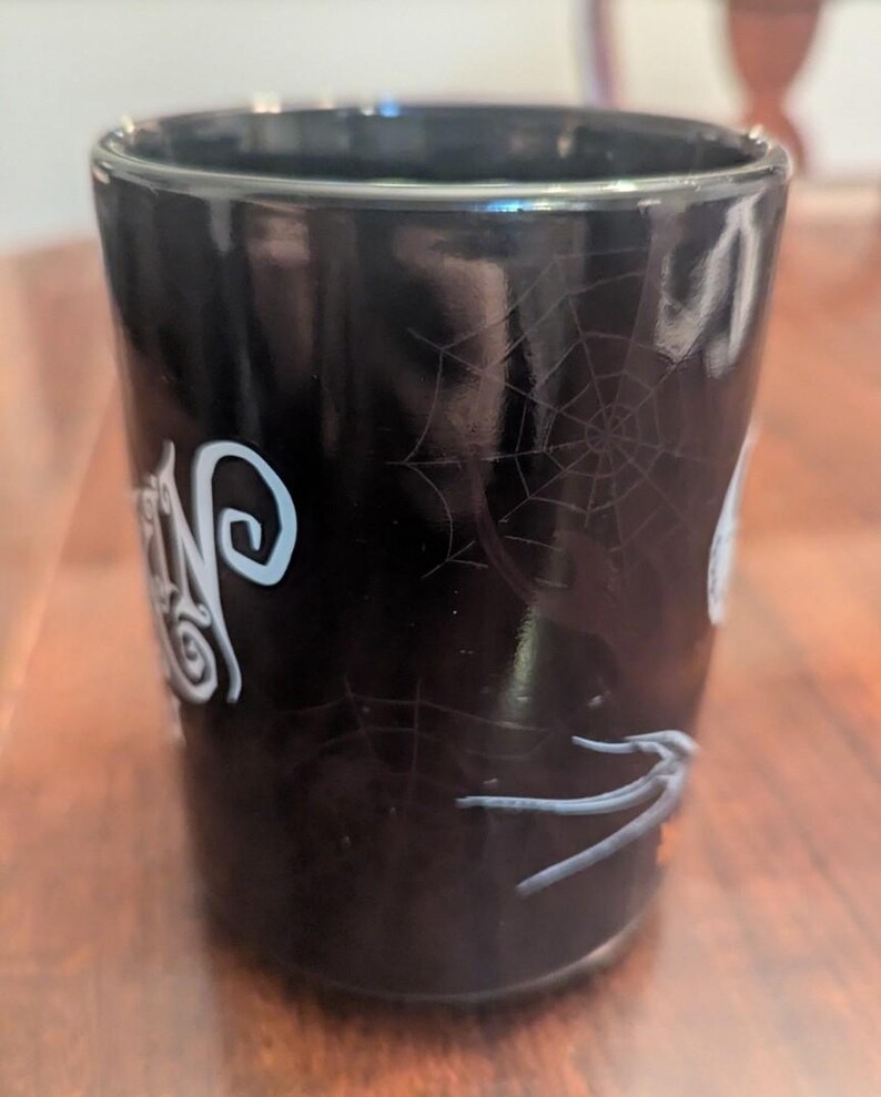 May include: A black cylindrical mug featuring a glossy finish with a white spiderweb pattern printed on the exterior. The design includes stylised white lettering and a grey spider graphic, creating a dark aesthetic on the smooth ceramic surface.