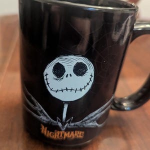 May include: A black ceramic mug featuring a white, round, stitched-face character graphic centred on the front. Above the character is a faint spiderweb design, and below is the orange text "NIGHTMARE". The mug has a glossy finish and a handle.