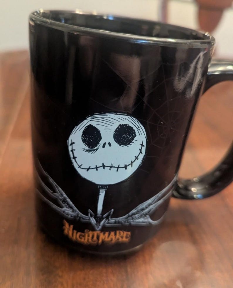 May include: A black ceramic mug featuring a white, round, stitched-face character graphic centred on the front. Above the character is a faint spiderweb design, and below is the orange text "NIGHTMARE". The mug has a glossy finish and a handle.