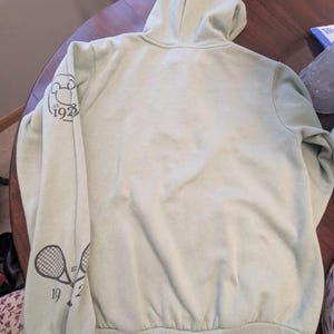 Puede incluir: Una sudadera con capucha verde claro con capucha y mangas largas. La manga izquierda presenta un gr&aacute;fico de raqueta de tenis y los n&uacute;meros "1923". La sudadera con capucha est&aacute; hecha de un material suave y tiene dobladillo y pu&ntilde;os acanalados.