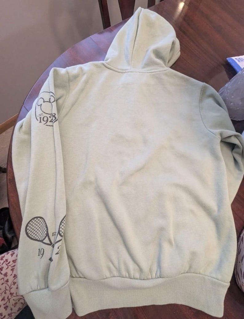 Puede incluir: Una sudadera con capucha verde claro con capucha y mangas largas. La manga izquierda presenta un gr&aacute;fico de raqueta de tenis y los n&uacute;meros "1923". La sudadera con capucha est&aacute; hecha de un material suave y tiene dobladillo y pu&ntilde;os acanalados.