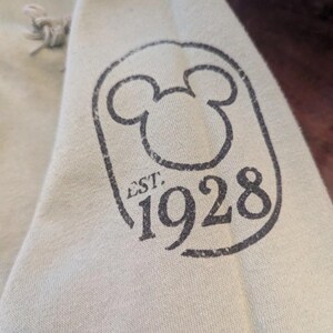 Puede incluir: Primer plano de una manga de sudadera con capucha verde claro con un gr&aacute;fico negro desgastado. El gr&aacute;fico presenta la ic&oacute;nica silueta de Mickey Mouse dentro de un marco rectangular, con el texto "EST. 1928" y el n&uacute;mero "1928" debajo.