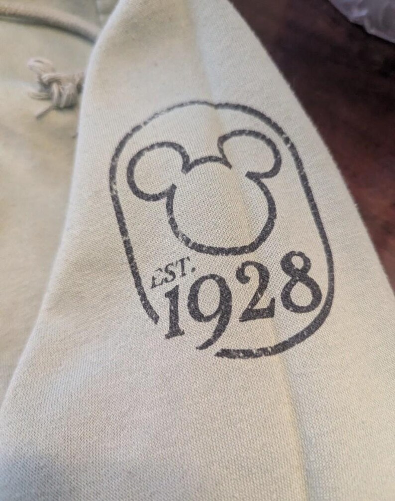 Puede incluir: Primer plano de una manga de sudadera con capucha verde claro con un gr&aacute;fico negro desgastado. El gr&aacute;fico presenta la ic&oacute;nica silueta de Mickey Mouse dentro de un marco rectangular, con el texto "EST. 1928" y el n&uacute;mero "1928" debajo.