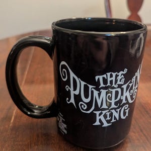 May include: A black ceramic mug featuring the white text The Pumpkin King in a stylised font. The mug includes a smaller white logo near the base that reads Disney The Nightmare Before Christmas. The handle is black and curved on the left side.