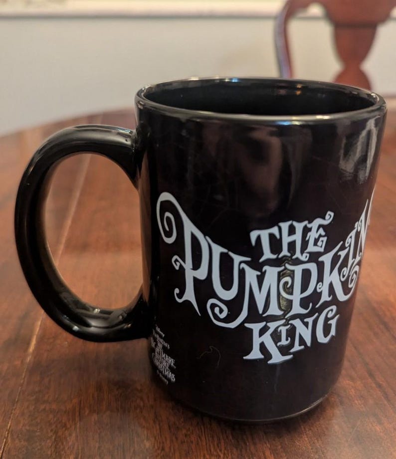 May include: A black ceramic mug featuring the white text The Pumpkin King in a stylised font. The mug includes a smaller white logo near the base that reads Disney The Nightmare Before Christmas. The handle is black and curved on the left side.