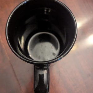 May include: A top-down view of a solid black ceramic mug. The mug features a smooth, glossy finish on both the interior and exterior surfaces. A single curved handle is attached to the side, and the circular opening reveals a deep, dark basin.