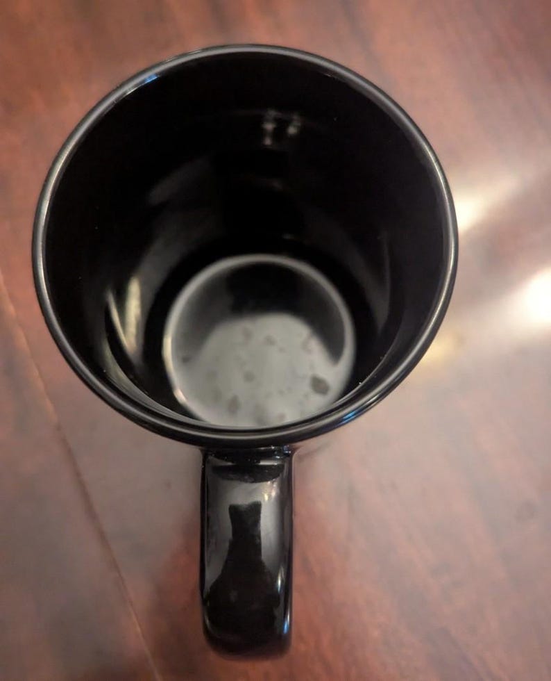 May include: A top-down view of a solid black ceramic mug. The mug features a smooth, glossy finish on both the interior and exterior surfaces. A single curved handle is attached to the side, and the circular opening reveals a deep, dark basin.
