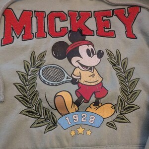 Puede incluir: Sudadera con capucha verde salvia con capucha con cord&oacute;n y bolsillo delantero. La parte delantera presenta la palabra "MICKEY" en letras rojas en bloque sobre una imagen de dibujos animados de Mickey Mouse jugando al tenis. La imagen incluye el a&ntilde;o "1928" y una corona de laurel.