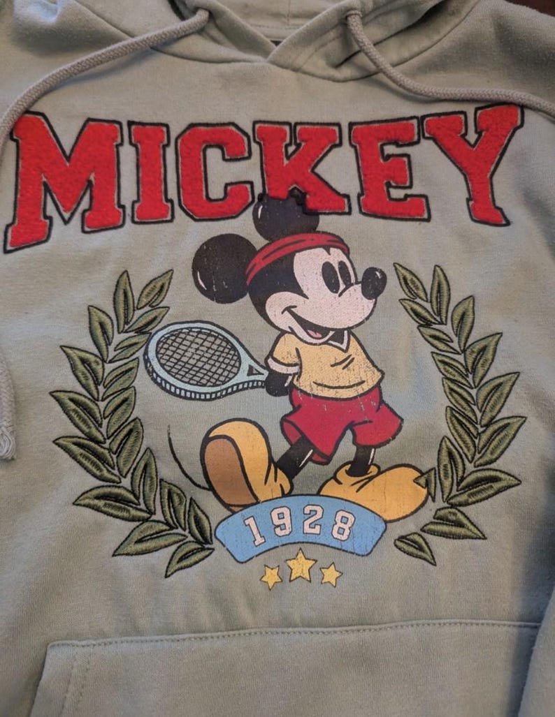 Puede incluir: Sudadera con capucha verde salvia con capucha con cord&oacute;n y bolsillo delantero. La parte delantera presenta la palabra "MICKEY" en letras rojas en bloque sobre una imagen de dibujos animados de Mickey Mouse jugando al tenis. La imagen incluye el a&ntilde;o "1928" y una corona de laurel.