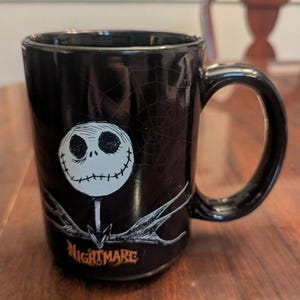 May include: A black ceramic mug featuring a white, round-faced character with stitched eyes and mouth. Below the character is a grey, skeletal bowtie and the orange text NIGHTMARE. A faint spiderweb pattern is visible on the black background.