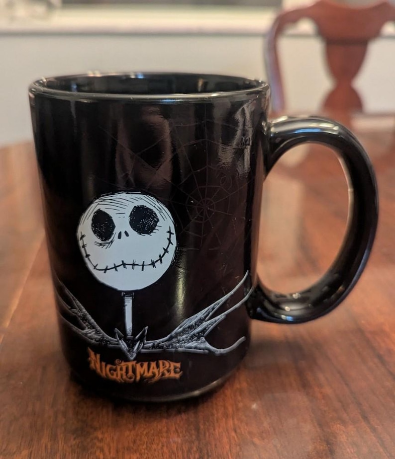 May include: A black ceramic mug featuring a white, round-faced character with stitched eyes and mouth. Below the character is a grey, skeletal bowtie and the orange text NIGHTMARE. A faint spiderweb pattern is visible on the black background.