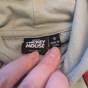 Puede incluir: Primer plano de una etiqueta de sudadera con capucha verde claro con el logotipo de "Mickey Mouse" y el texto "Made in China". La etiqueta tambi&eacute;n indica la talla "S". El logotipo de Disney tambi&eacute;n es visible.