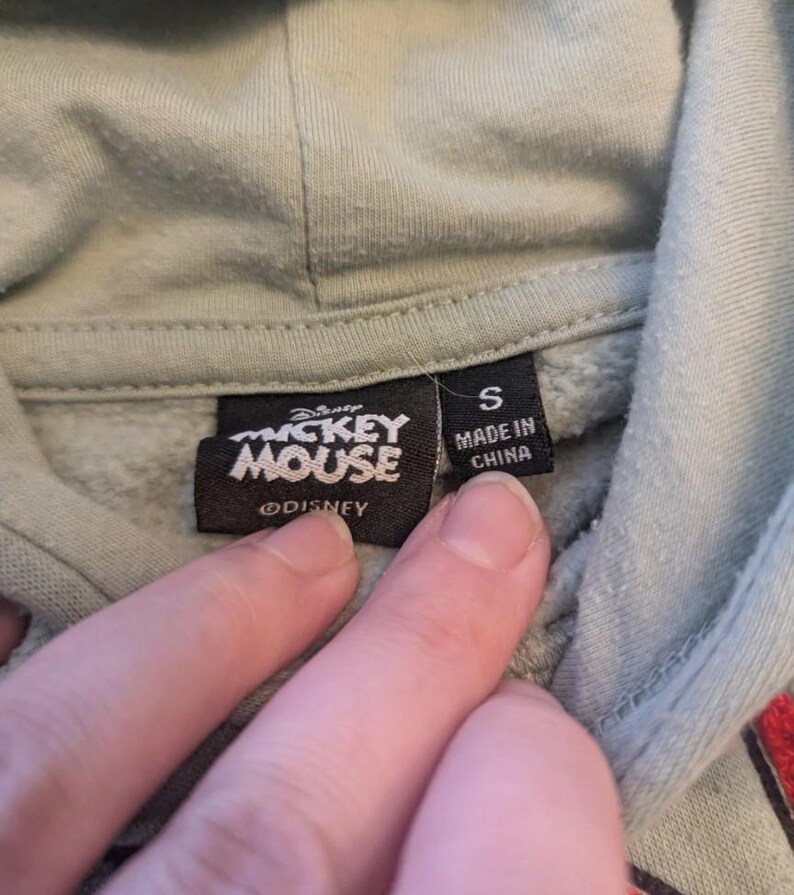 Puede incluir: Primer plano de una etiqueta de sudadera con capucha verde claro con el logotipo de "Mickey Mouse" y el texto "Made in China". La etiqueta tambi&eacute;n indica la talla "S". El logotipo de Disney tambi&eacute;n es visible.