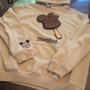 May include: A beige hooded sweatshirt featuring a large, textured brown Mickey Mouse-shaped ice cream bar graphic on the chest. The text Disneyland Resort is embroidered below the graphic, and a small Mickey Mouse face is on the right sleeve.