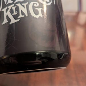 May include: A black ceramic mug featuring the white text PUMPKIN KING printed in a stylised font. The surface of the mug has a glossy finish with a subtle spiderweb graphic visible on the side. The base of the mug is rounded and smooth.