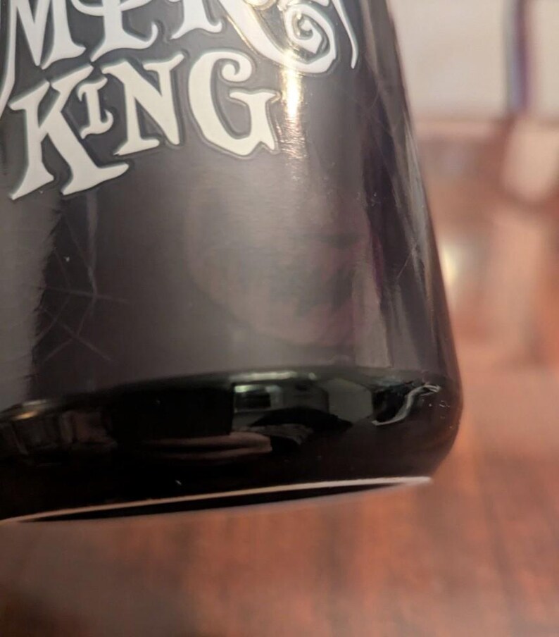 May include: A black ceramic mug featuring the white text PUMPKIN KING printed in a stylised font. The surface of the mug has a glossy finish with a subtle spiderweb graphic visible on the side. The base of the mug is rounded and smooth.