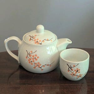May include: A white ceramic teapot with a matching teacup, both featuring a delicate hand-painted design of brown branches with small orange blossoms. The teapot has a rounded body, a curved handle, a short spout, and a lid with a knob handle.