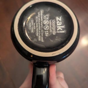 May include: The underside of a black ceramic mug featuring white printed text that reads zak! designs, 13+, and instructions to hand wash gently, do not soak, and not to microwave. The base also includes the date 03/2023 and China 15294-1.