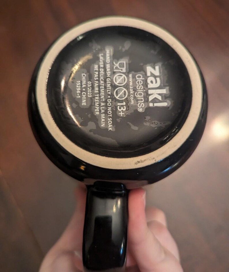 May include: The underside of a black ceramic mug featuring white printed text that reads zak! designs, 13+, and instructions to hand wash gently, do not soak, and not to microwave. The base also includes the date 03/2023 and China 15294-1.