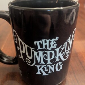 May include: A black ceramic mug featuring the text THE PUMPKIN KING printed in a stylised light blue font on the front. The mug has a smooth, glossy finish and a curved handle on the left side. A faint, subtle spiderweb pattern is visible in the background.